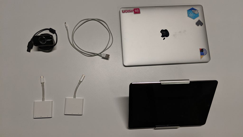 Top view of a MacBook pro laptop, an ipad table, two adapters, an ipad charging cable and a USB camera