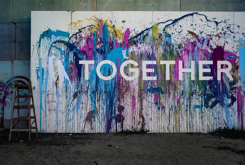 A painted canvas stands against a wall. It has a variety of paint splashes in blue, pink, yellow, purple. The middle of the canvas in white letters has the word: “Together.”