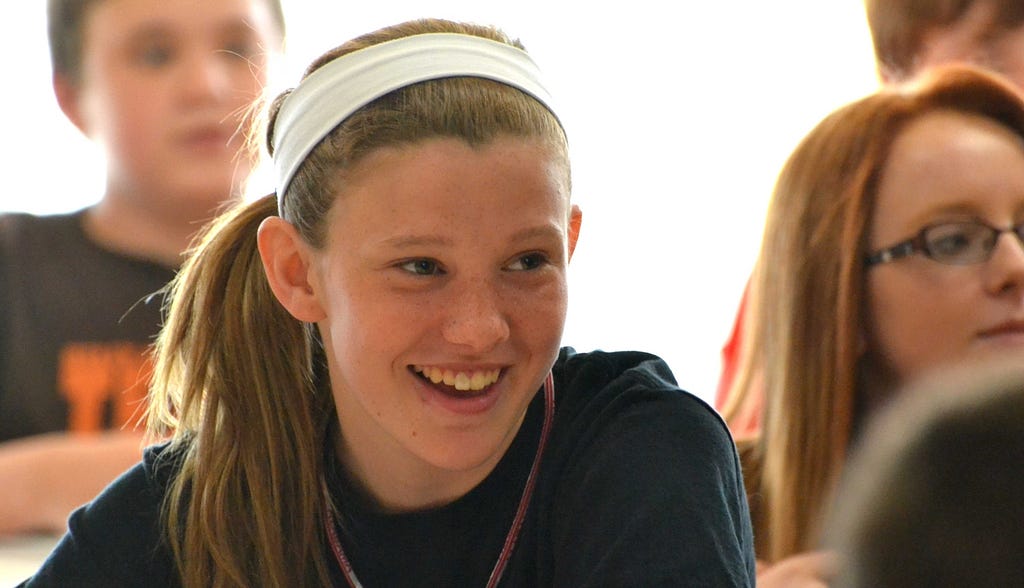 A student wearing a white headband and a pony tail smiles.