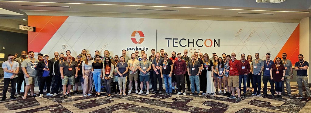 A large group of Paylocity employees stand in front of a wall showing the Paylocity TechCon 2023 logo.