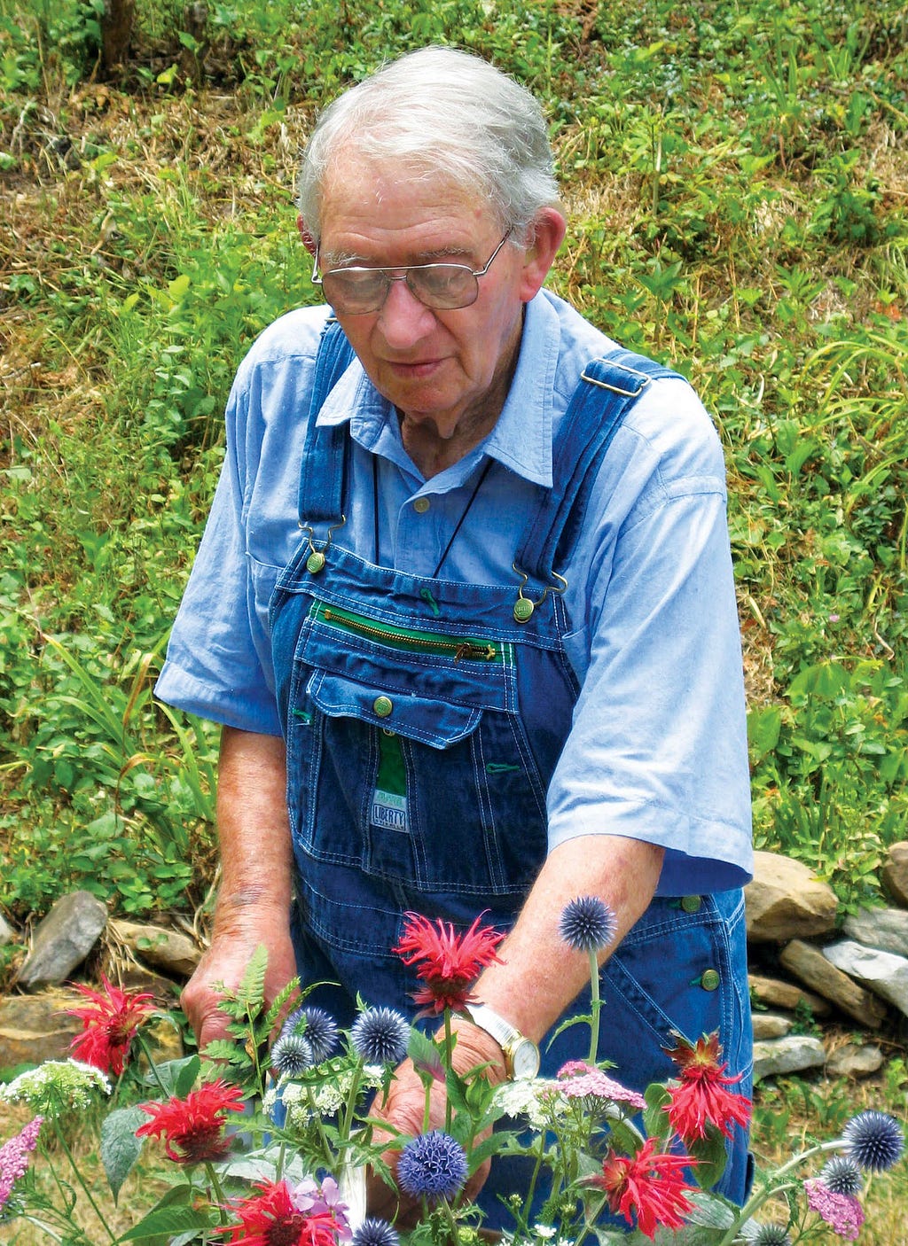 elderly white man in overalls and glasses touches a colorful flower arrangement