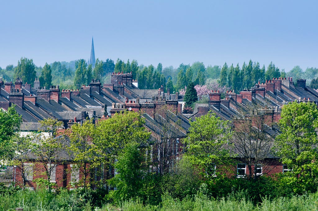 Terraces of houses near the city centre of Stoke on Trent, between lines of green-leaved trees. In the background, a church steeple rises above the treeline into a cloudless sky.