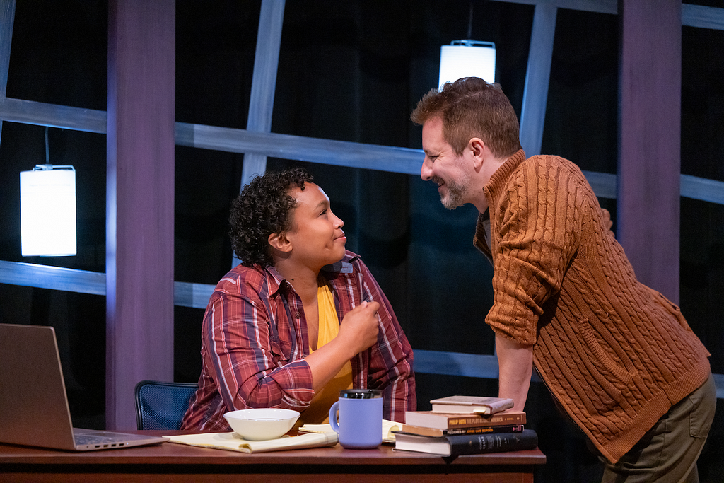 A woman in a plaid shirt sits at a cluttered desk with an open laptop. She smiles at a man, also smiling, who leans on the desk.