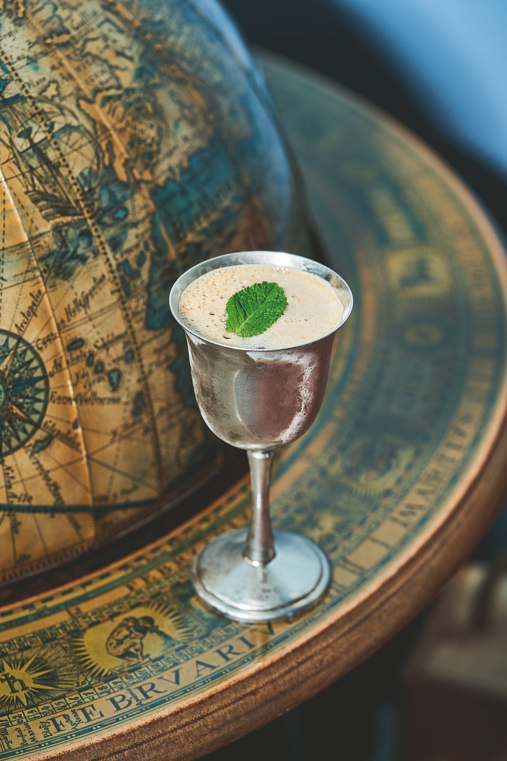 a metal goblet with a foam-topped drink and leaf on top with a globe in the background