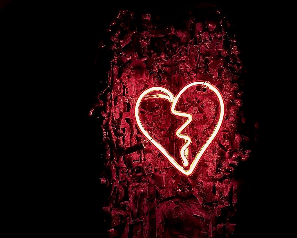 A neon broken heart sign lights up the background, filled with gears and industrial material, red.