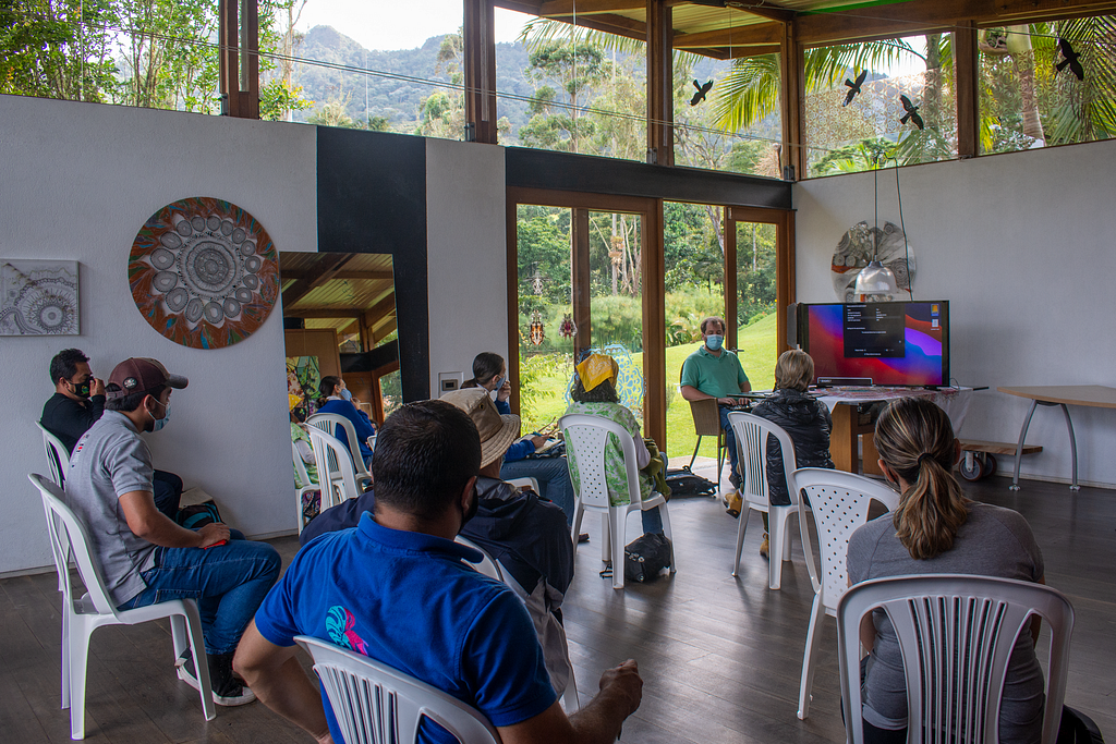 A group of people sitting in white outdoor chairs attends one of Juan Pablo’s workshops about the birdwatching trail as he sits at the head of the class facing the group.
