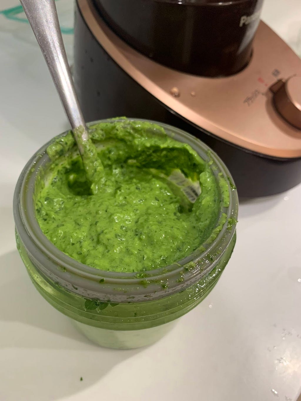 A jar filled with homemade parsley paste, made with parsley, garlic, and olive oil. A spoon is resting in the jar, and a kitchen appliance is visible in the background.