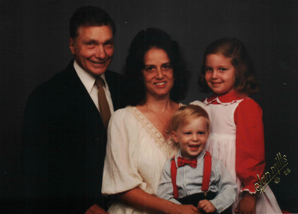 A family of four, left to right: a father wearing a tie, a mother wearing a white shirt with black hair, and a daughter with a red and white dress. In front of them is a little boy wearing a light blue shirt and red suspenders and a bow tie. In the lower right, there is a watermark noting that it was taken by Olan Mills in 1988.