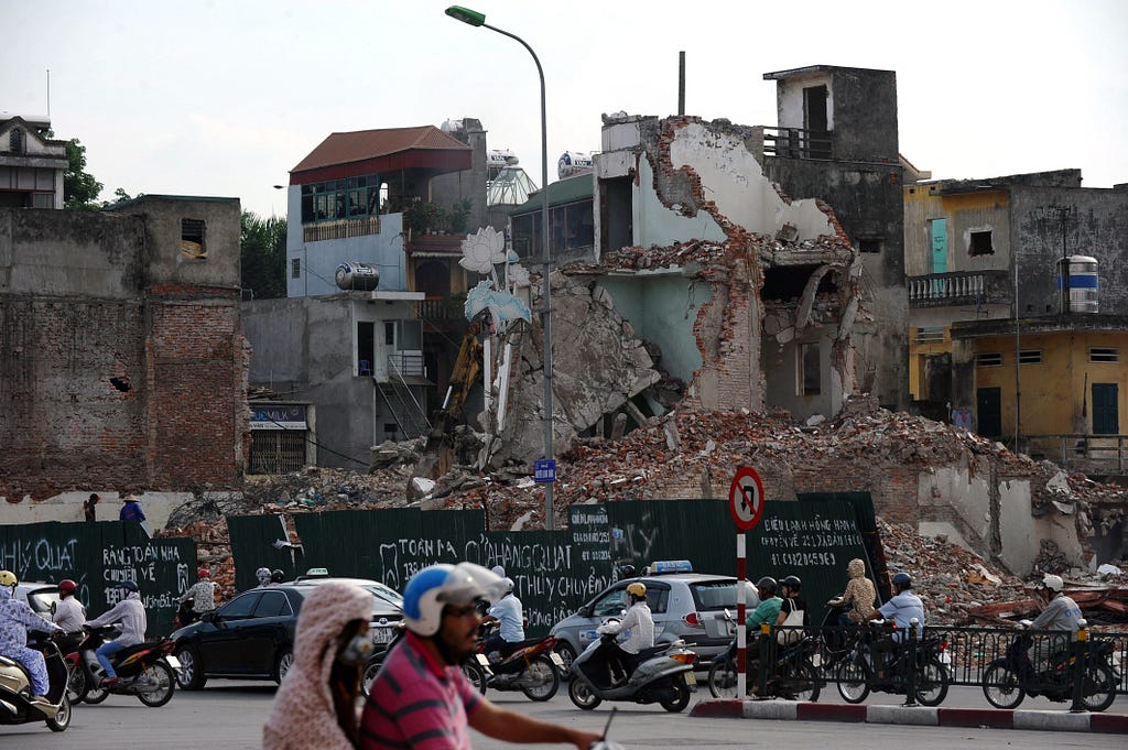 Car and motorcycle traffic passes by a decaying urban structure.