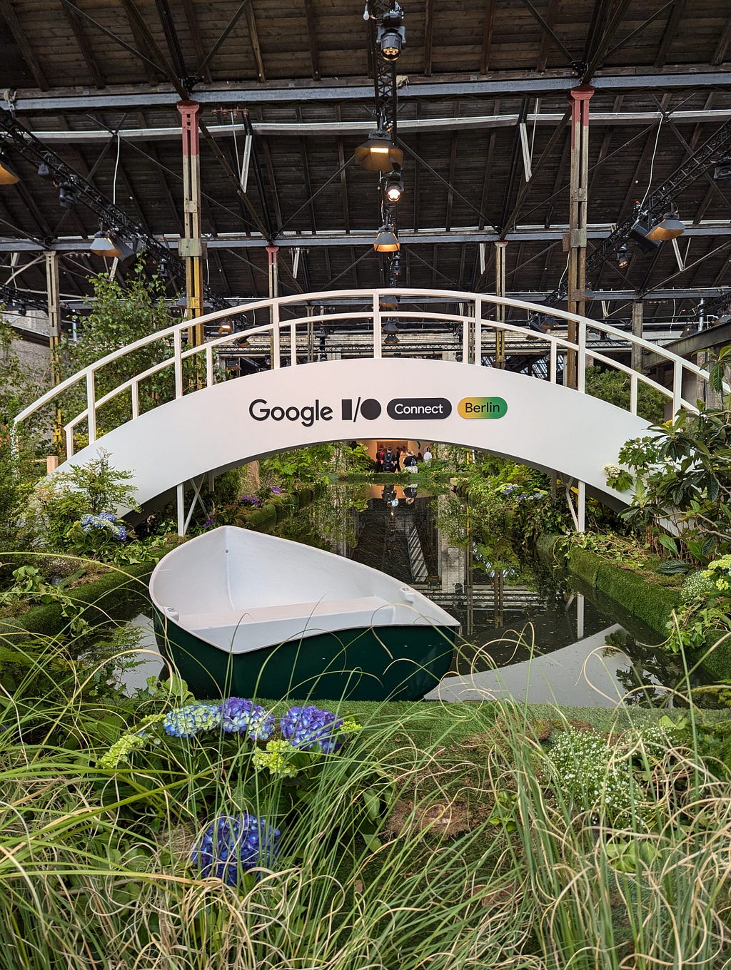 An artificial indoors river with a boat in the foreground and lush green riverbanks and a white bridge with the Google I/O Connect logo on spanning it