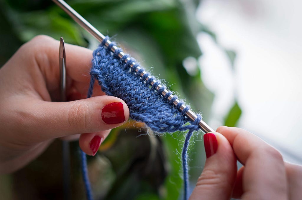 White hands with red nail polish knitting blue yarn.