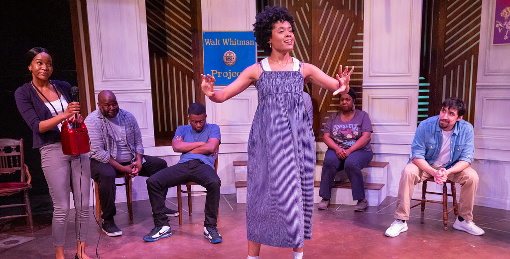 A group of people sit in chairs listening to a person with brown skin and dark brown hair in a blue sleeveless dress give a speech. A person with brown skin and black hair holds a small microphone and speaker on the left side.