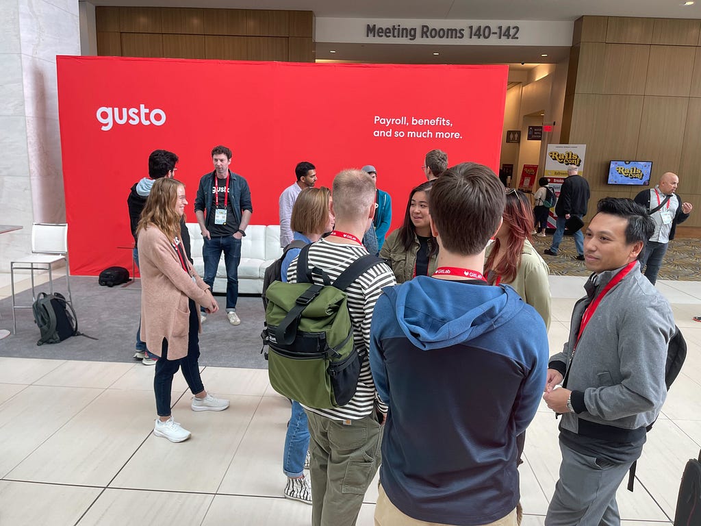 Several people gathered and chatting in front of a guava-colored Gusto backdrop at RailsConf.
