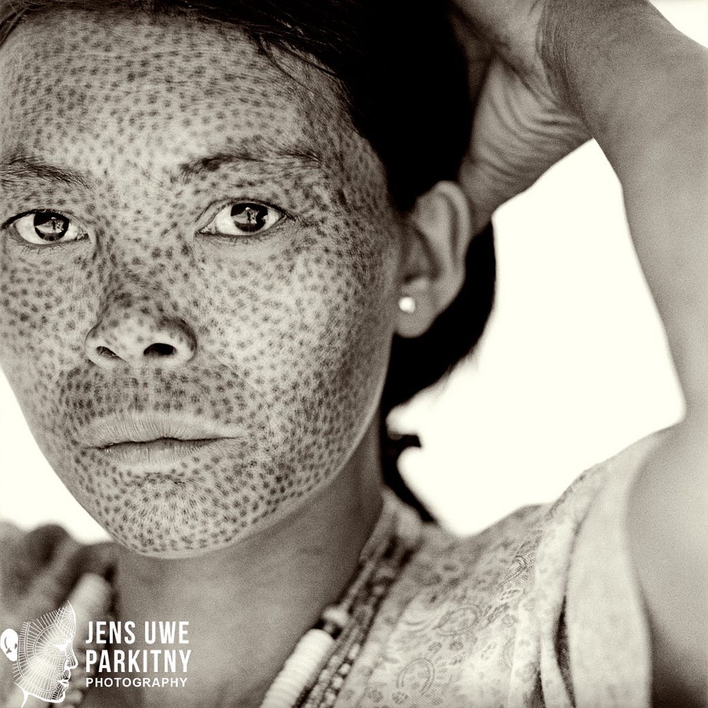 A portrait of a Matu Chin women with her groups visual signature tattooed all over her face: dots, that look like freckles.