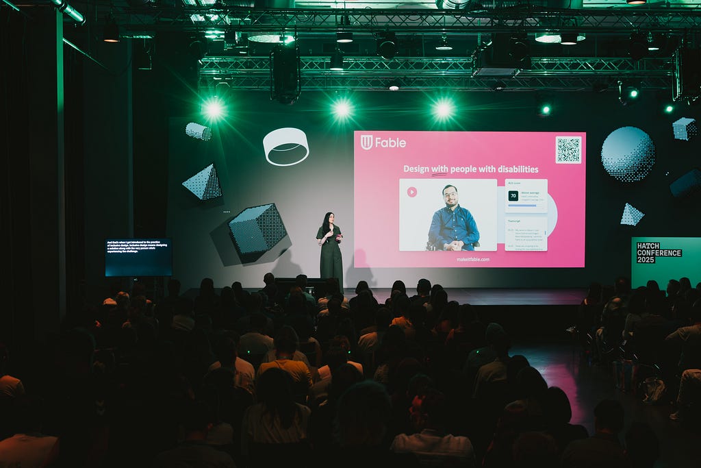 A person in a long green dress standing on a well-lit stage and giving a presentation to a dark room full of seated people, with a graphic on a large screen behind the speaker that reads “Design with people with disabilities”.