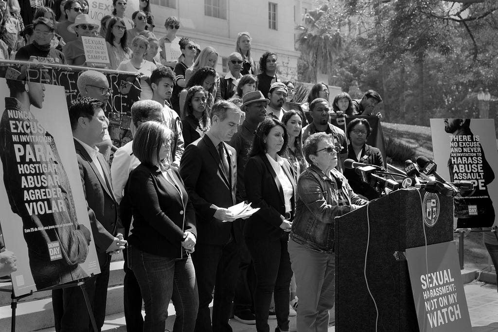 A woman in all denim stands speaking behind a podium. Behind her, a large crowd stands in solidarity, many of which are also wearing denim.