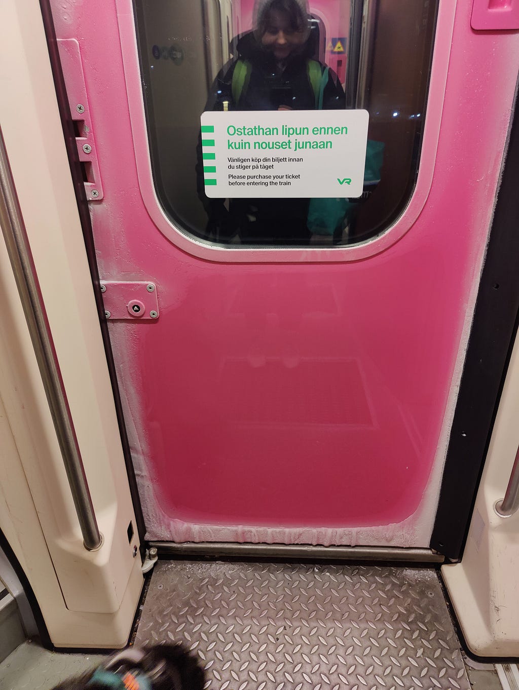 A photo of a bright pink train door, taken from the inside. At the top of the door is a rectangular window with rounded corners and a white notice inside. Around the frame of the door is a thick layer of frost creeping over, turning pink to white.