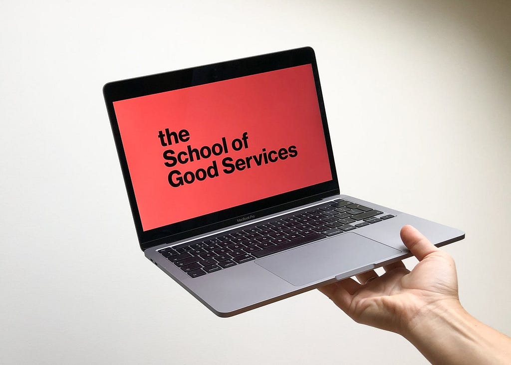 a hand holds a laptop with a slide on the screen that reads in big bold letters ‘The School of Good Services’