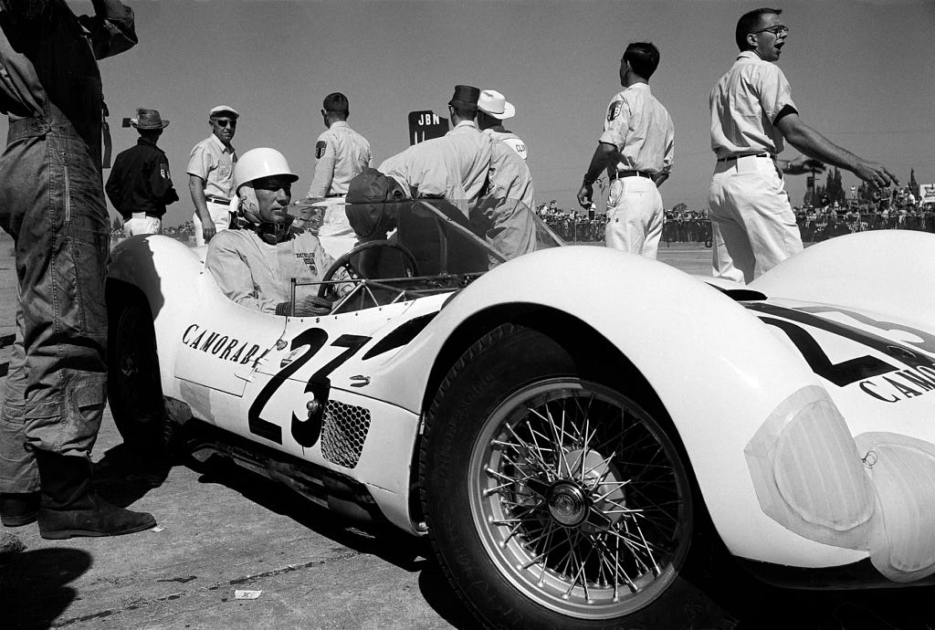 A serious-looking man in a white helmet sits in an old-fashioned open-top racecar with the words “Camorare 23” emblazoned on the side.