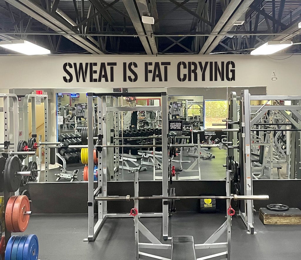 a photo of a gym with weight racks. The wall has printing on it in large black letters “SWEAT IS FAT CRYING”