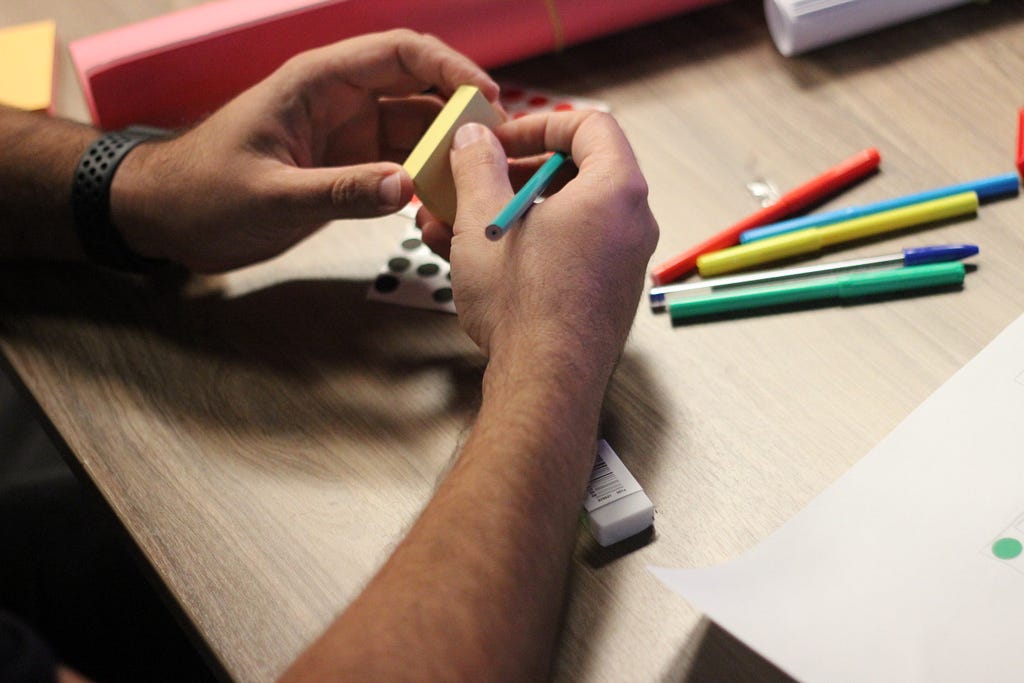 Foco em mãos masculinas, apoiadas em uma mesa de madeira, segurando um bloco de post-its e canetas coloridas sob a mesa.