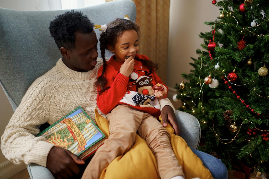Father and daughter sit with a book by the Christmas tree