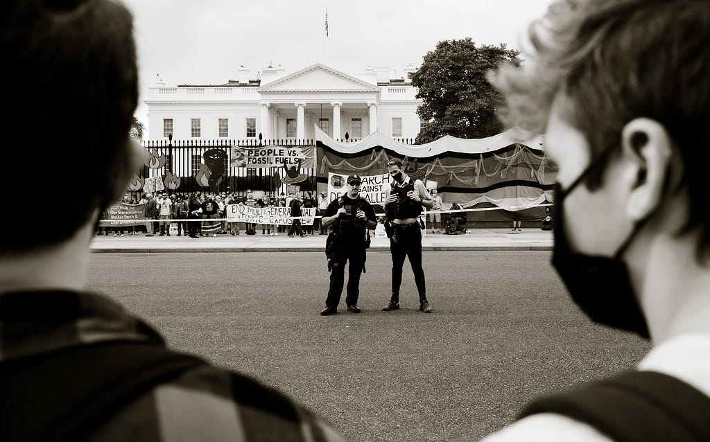 In the foreground two white people wearing respirators look at two cops, one wearing a respirator, both dressed in riot gear. Behind them are stalls and banners from a climate protest.