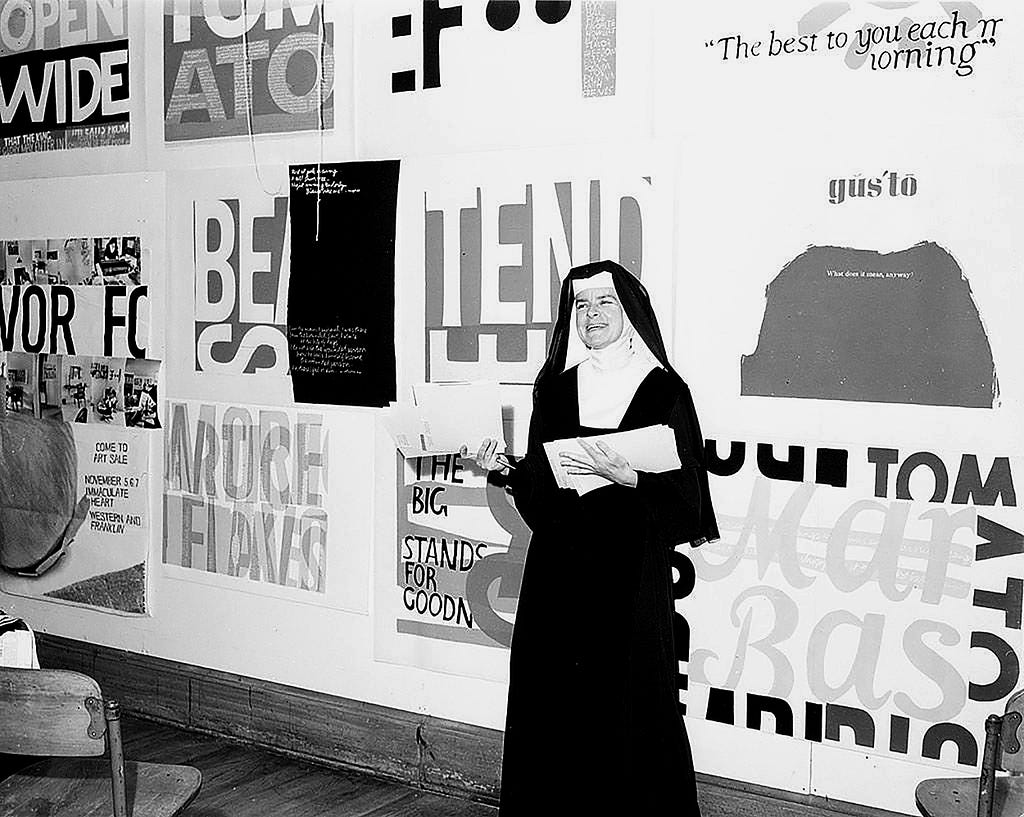 Photography of Sister Corita Kent stands in front of her work, including for Eleanor, at Immaculate Heart College in 1964.