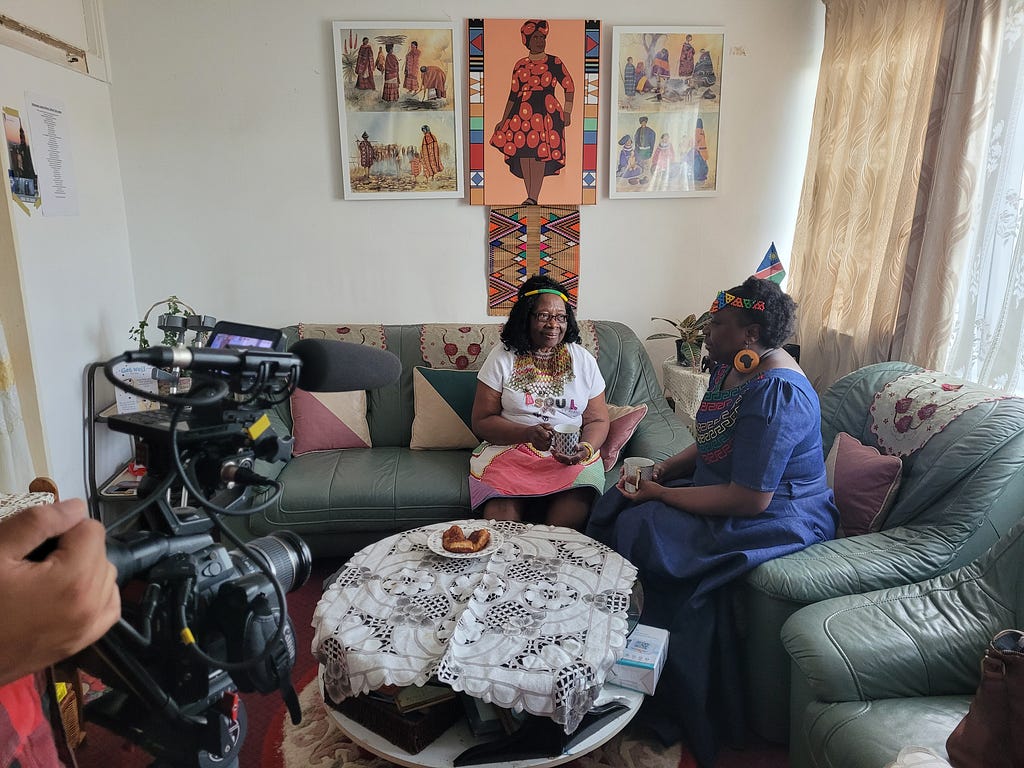 Thabani, an older woman, sits with her daughter Khwezi on sofas in their home. They’re talking to each other while a TV camera can be seen filming their conversation.