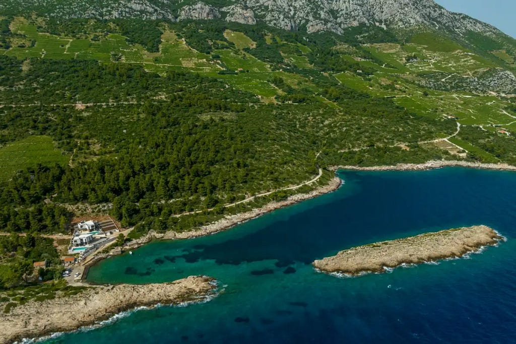 Aerial view of a picturesque coastal landscape featuring lush green hillsides and terraced vineyards. The shoreline is dotted with rocky outcrops and a small cove with turquoise waters. At the edge of the water, there are a few buildings nestled among the trees, suggesting a secluded and serene retreat. In the distance, the rugged terrain of the mountains creates a stunning backdrop against the deep blue sea.