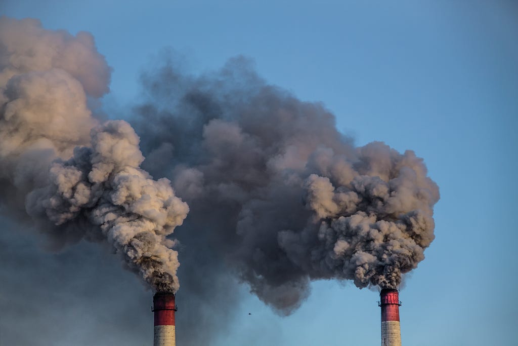 Two red and white smoke stacks releasing dark smoke into the air. Photo by kapichka/Adobe Stock