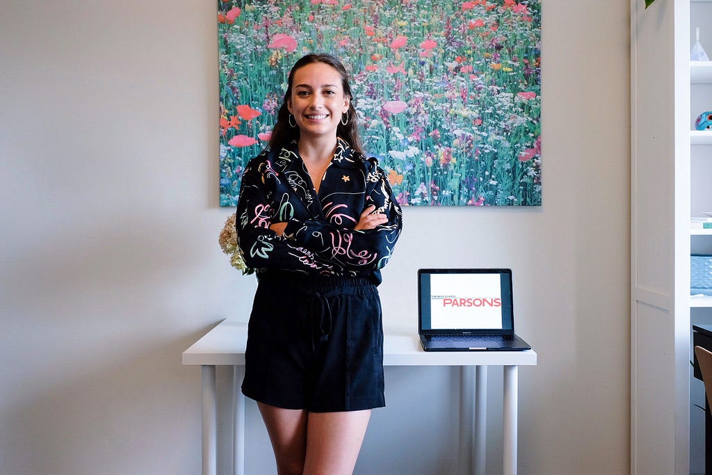 Lina Alvarez standing in front of a desk with arms crossed. On a table behind her is a laptop with the Parsons School of Design Logo on the screen