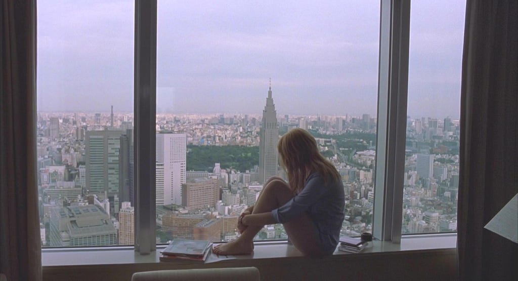 A woman sitting in front of the window, gazing at the panoramic view of the city and the clock tower in the background at dusk.