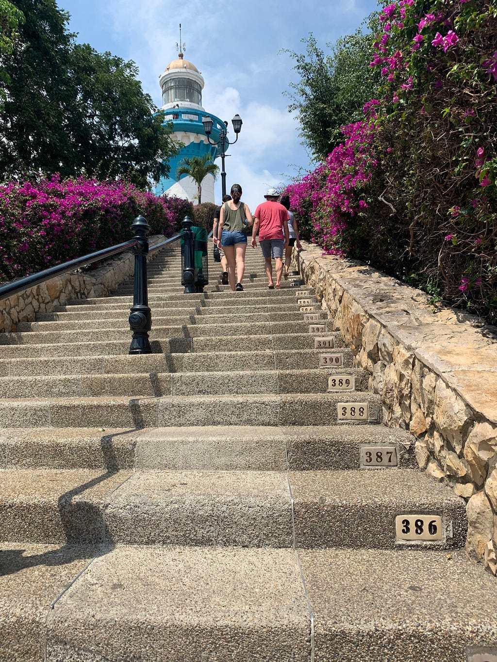 The numbered steps of Las Peñas are visible. Four people walk up the steps. Purple flowers are on either side of the stairs.
