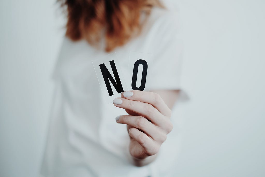 Person with red hair, photographed from the shoulders down, in a white t-shirt on a white backdrop, holding out the letters N and O to make “NO” in their delicate hand. They have white nail polish on and are holding out their left arm.
