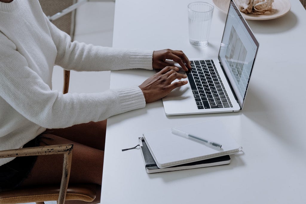 Mulher digitando em um notebook. Em cima da mesa há também um copo, um prato e um caderno ao lado do notebook