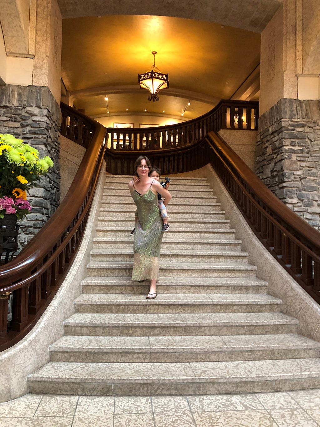 The author walking down some elegant stairs dressed in a sparkling sheath dress carrying a small child piggyback