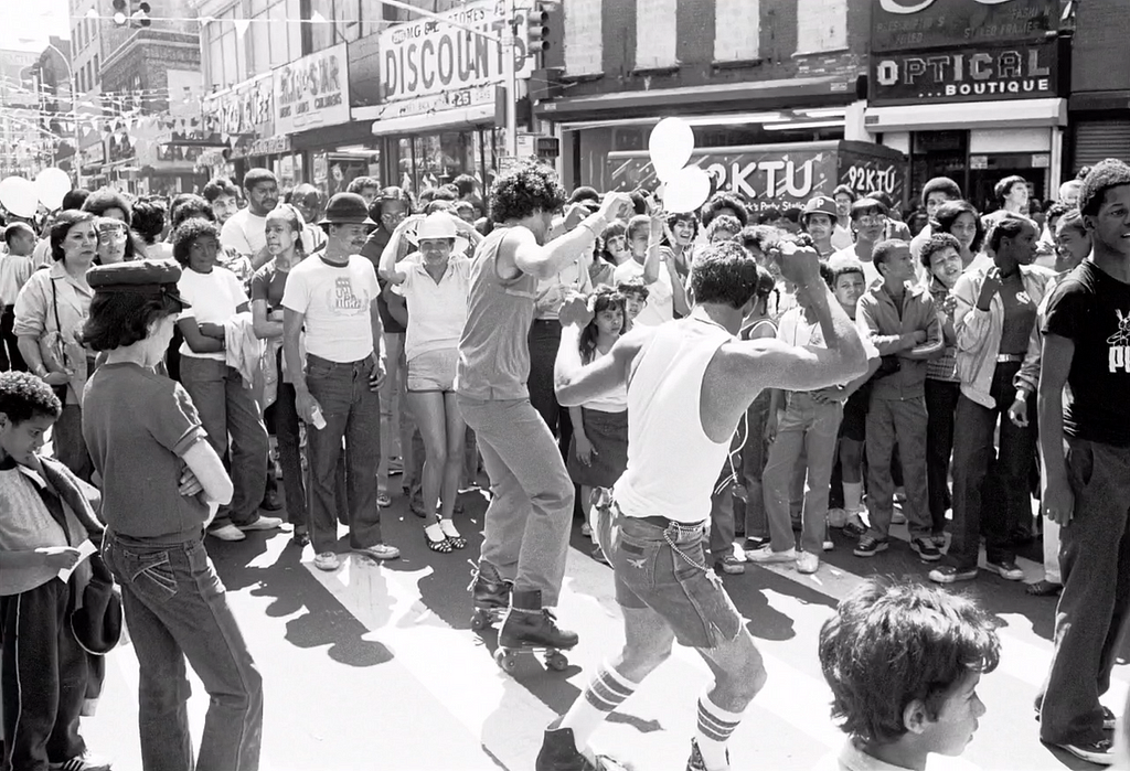 Dancers on rollerskates are dancing at a block party next to a contemplative crowd.