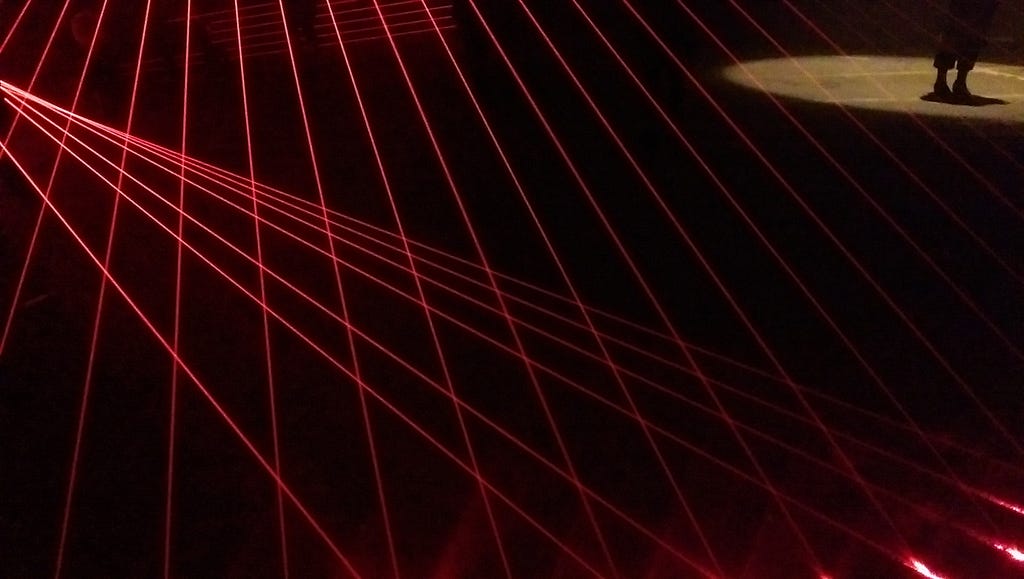 Red laser beams criss-cross the darkness. In the top right corner, a person stands in a pool of light.