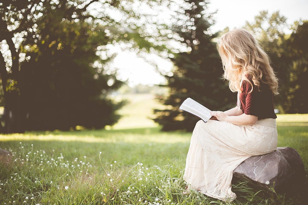 A blonde woman sitting on a rock in a park, reading a book