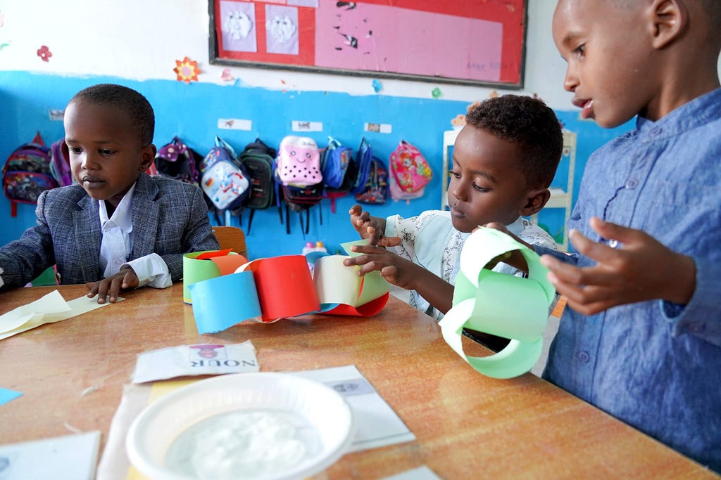 Three preschool boys at use blue, red, and green paper to make a toy at their desk. Tiny kid’s backpacks hand on a wall behind them.