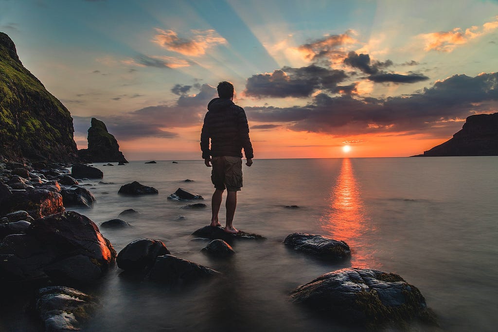 Photo of a man standing on a rock in the water. He’s wearing shorts and a long-sleeve jacket. He’s facing away from the camera and looking at a pink orange sunrise, peaking on the horizon at the distant water’s edge. The sun’s rays shine up and out, piercing the grey and white clouds in the sky. The edges of the photo show rocks and mountain in the dark foreground on the left, near the man. There’s a mountain or land in the far background on the right.