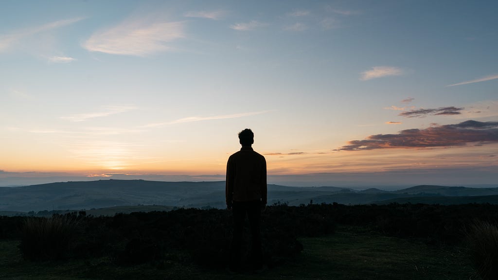 Man standing alone in a field with the sun setting in the background.
