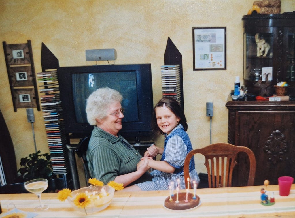 Family photo of the author as a young child sitting on her grandmother’s lap. Both are smiling at a table where a birthday cake with lit candles awaits.