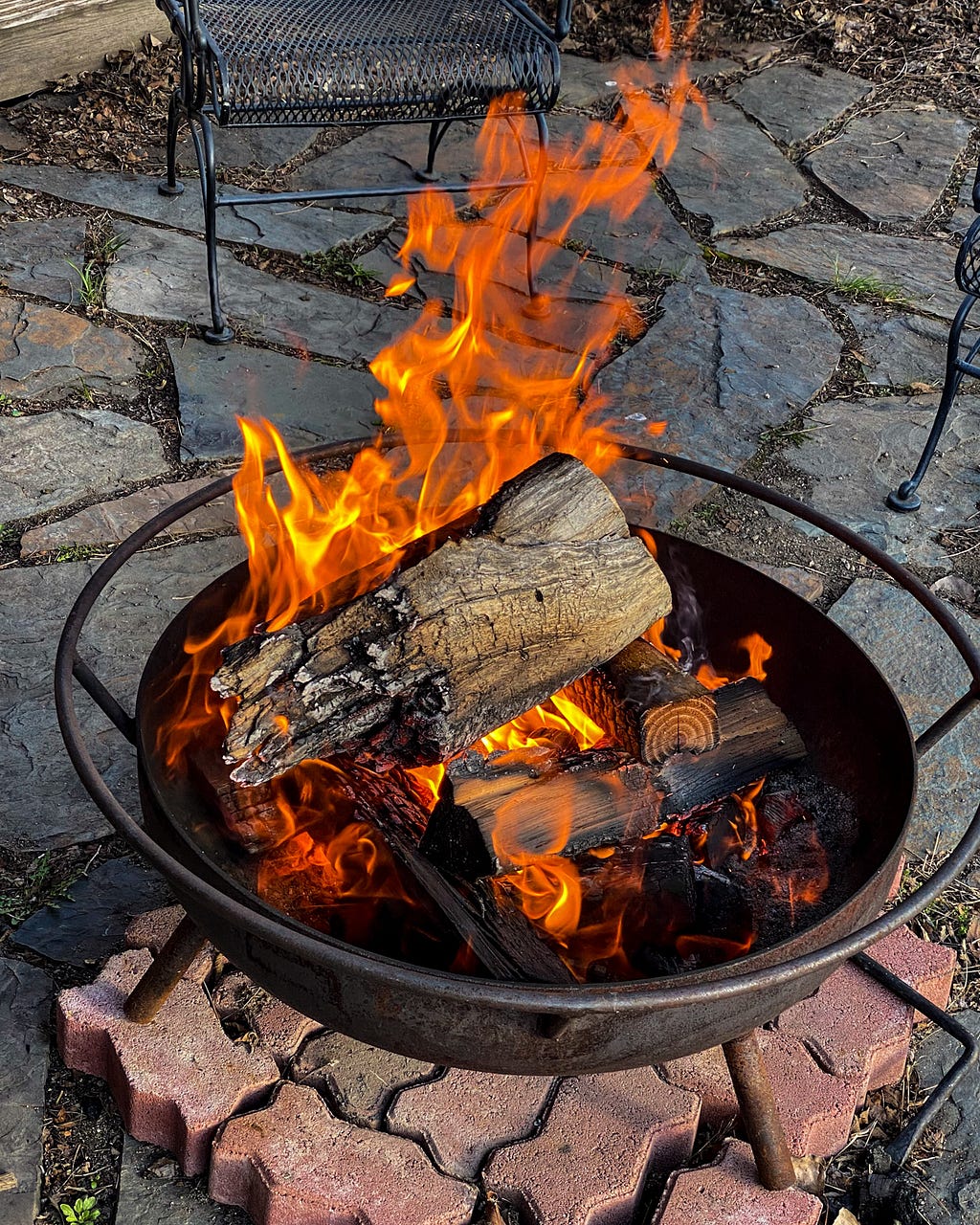 A picture of a wood fire in a metal, standalone fire place on an outdoor, flagstone patio.