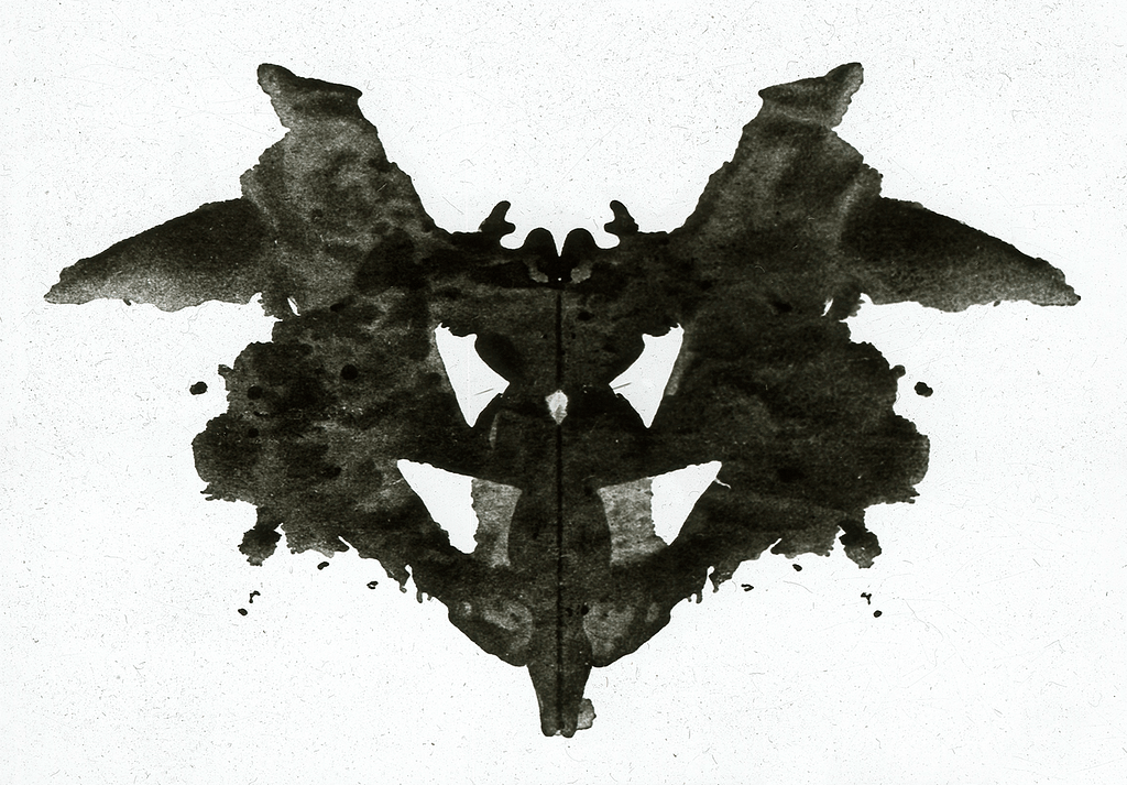 A photograph of a Rorschach blot. It’s black ink smeared on a white piece of paper that’s been folded in half to smoosh the ink into an abstract shape. When they folded the paper with wet ink on it, it created a mirrored pattern of the ink as it spread out across the paper.