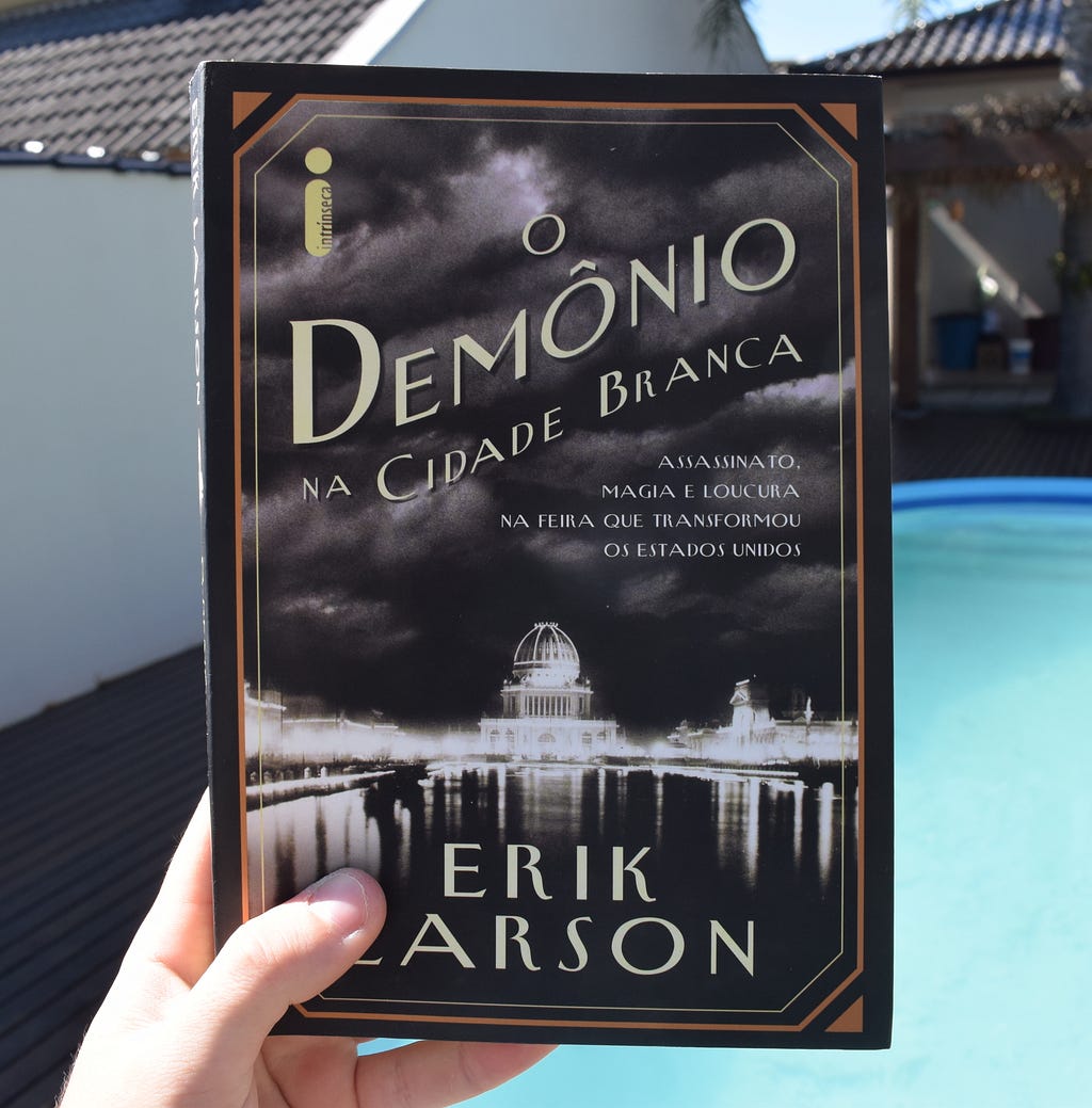 Uma mão segura um exemplar do livro O Demônio na Cidade Branca, de Erik Larson; em segundo plano, há uma casa e uma piscina.