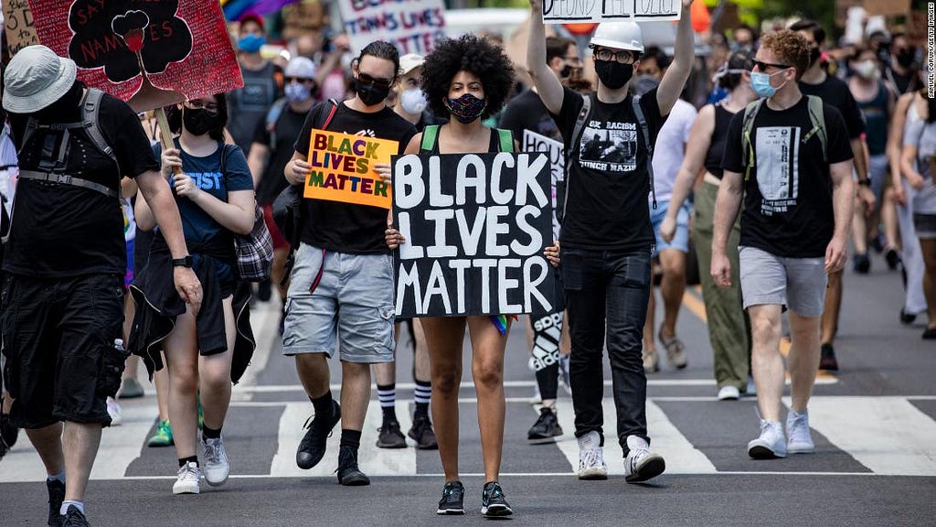 A protest march with a young African-American woman at the center in a face mask. Her sign reads, “Black Lives Matter.”