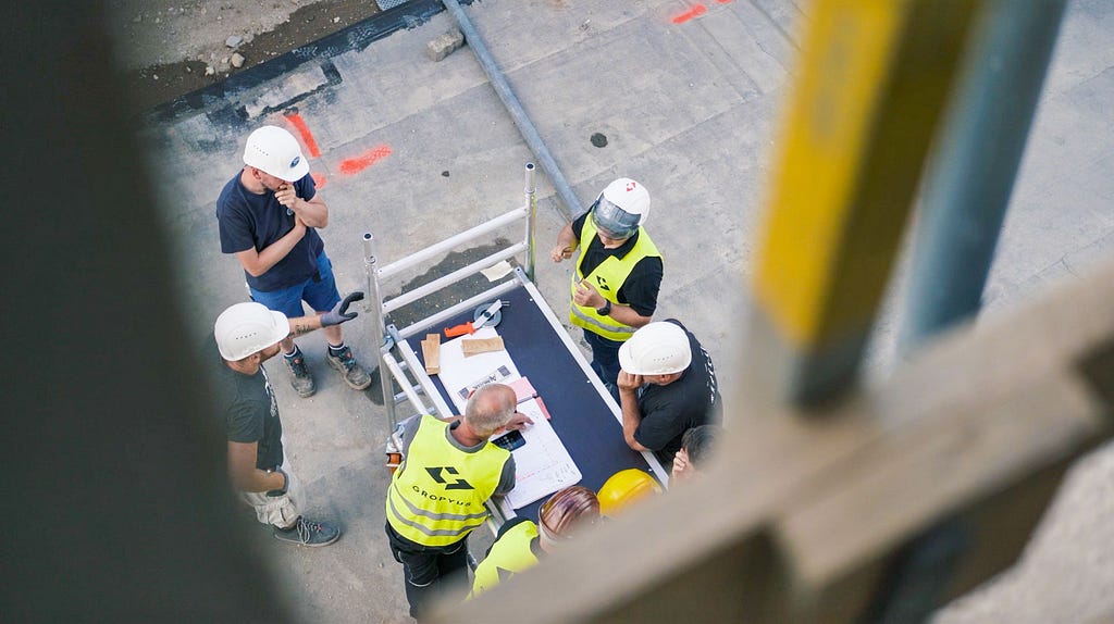 Six men, some wearing neon yellow vests, having a conversation around a table on a building site.