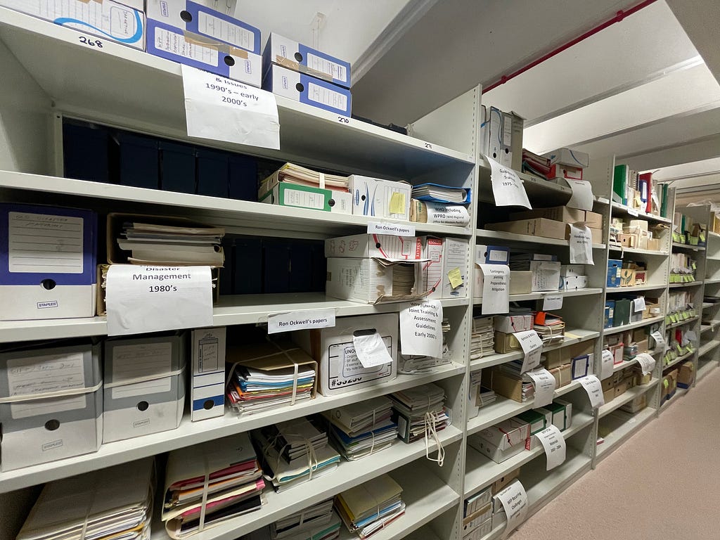 Four shelves full of boxes of documents and bound piles of documents. Many of the boxes and piles have sheets of paper denoting their contents. At the very right of the image is a fifth, emptier shelf.